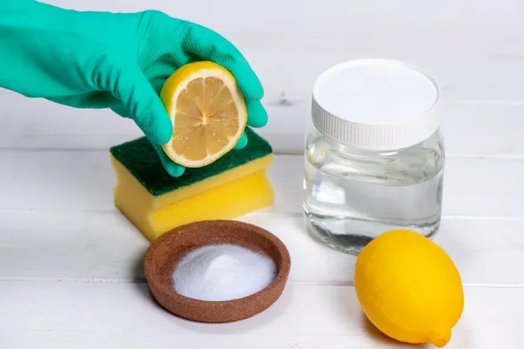un guanto tiene una fetta di limone sopra una spugna, vicino c'è una ciotola di bicarbonato e barattolo d'aceto 